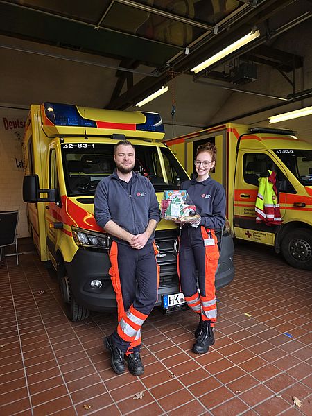 Zwei Mitarbeitende der DRK Bereitschaft stehen vor dem Rettungswagen und nehmen die Weihnachtsgeschenke der Kinder entgegen.